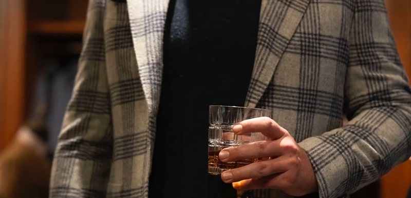 A person in a plaid jacket holds a glass of amber liquid.